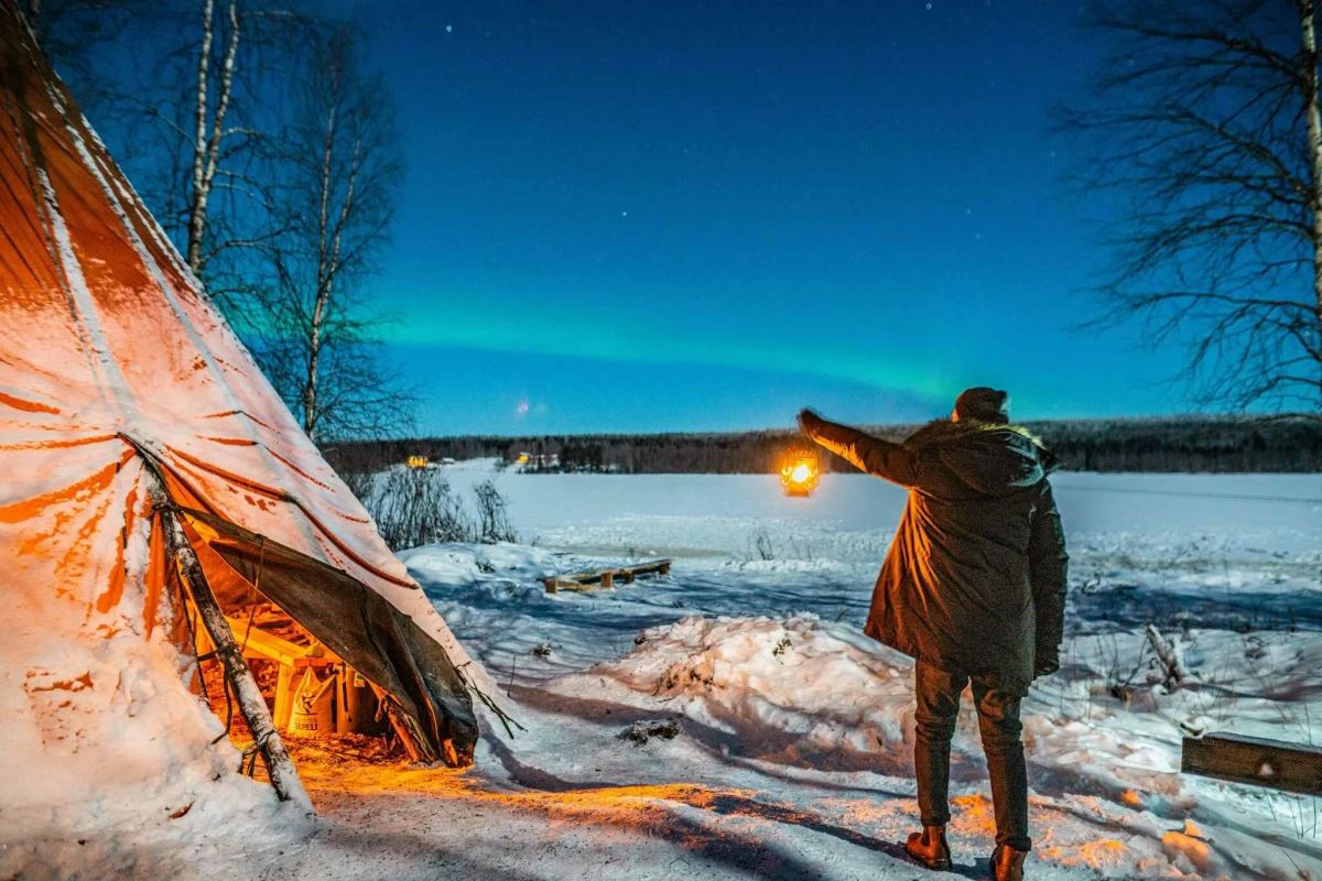 Traveller holding a lantern beside a wilderness tent beneath the Northern Lights